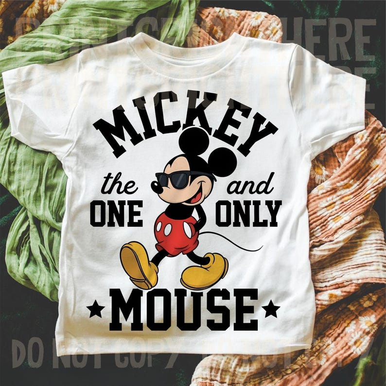 May include: White t-shirt with a Mickey Mouse graphic wearing sunglasses. The text reads "Mickey the One and Only Mouse." Mickey is in red shorts, yellow shoes, and black gloves. The design includes black stars. The t-shirt is a children's garment.