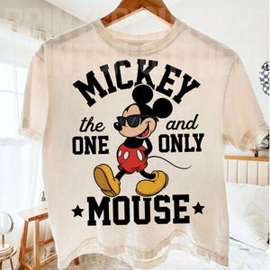 May include: Cream-coloured t-shirt featuring a graphic of Mickey Mouse wearing sunglasses, with the text "Mickey the One and Only Mouse" in black. Mickey is depicted in his classic red shorts, yellow shoes, and white gloves.