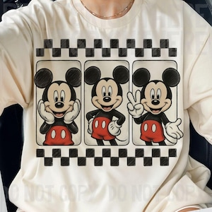 May include: Cream-colored t-shirt featuring a graphic of Mickey Mouse in three different poses. Mickey is wearing red shorts, white gloves, and black shoes. The design is framed by a black and white checkered pattern.