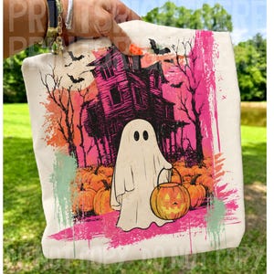 May include: A cream-coloured tote bag featuring a graphic print of a white ghost holding a jack-o'-lantern. The background shows a dark, stylised haunted house, bare trees, and flying bats in shades of vibrant pink, orange, and black ink.
