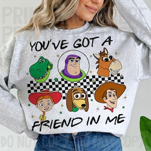 Può includere: Una felpa grigio chiaro con personaggi del film Toy Story. Il testo "YOU'VE GOT A FRIEND IN ME" è stampato in nero. Il design include immagini di Buzz Lightyear, Woody e altri personaggi del film d'animazione.