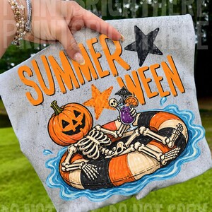 May include: A light gray t-shirt featuring a graphic of a skeleton with a pumpkin head floating in a black and orange striped inner tube on blue water. The skeleton holds a purple cocktail with an umbrella. The text reads SUMMER WEEN.