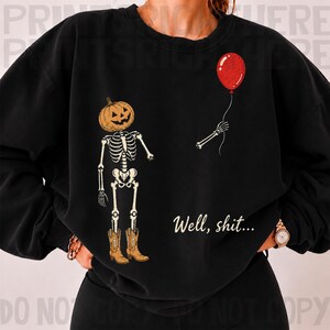Op de afbeelding: Een zwart sweatshirt met ronde hals en een afbeelding van een skelet met een pompoenhoofd en bruine cowboylaarzen. Het skelet houdt een touwtje vast dat verbonden is met een rode ballon. De tekst "Well, shit..." staat in het wit eronder.