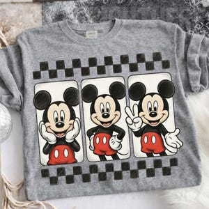 May include: A gray sweatshirt featuring three panels with Mickey Mouse illustrations. Each panel shows Mickey in a different pose, with a black and white checkered pattern above and below the panels. Mickey is wearing red shorts and white gloves.