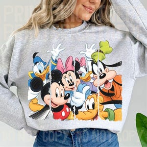 May include: A light gray sweatshirt featuring a colorful graphic of Disney characters including Mickey Mouse, Minnie Mouse, Donald Duck, Daisy Duck, and Goofy. The graphic has a distressed, vintage look.
