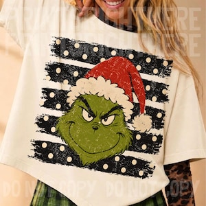 May include: A cream-colored t-shirt featuring a graphic of a green character with a furrowed brow wearing a red Santa hat. The design is set against a background of horizontal black brushstroke stripes with white polka dots throughout the print.