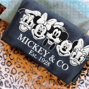 May include: A dark blue rectangular pillow featuring embroidered images of Mickey Mouse, Minnie Mouse, Donald Duck, Daisy Duck, and Goofy. The text "MICKEY & CO Est. 1928" is embroidered in white.