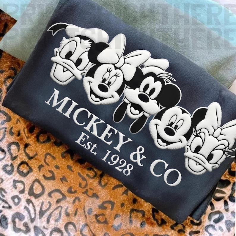 May include: A dark blue rectangular pillow featuring embroidered images of Mickey Mouse, Minnie Mouse, Donald Duck, Daisy Duck, and Goofy. The text "MICKEY & CO Est. 1928" is embroidered in white.