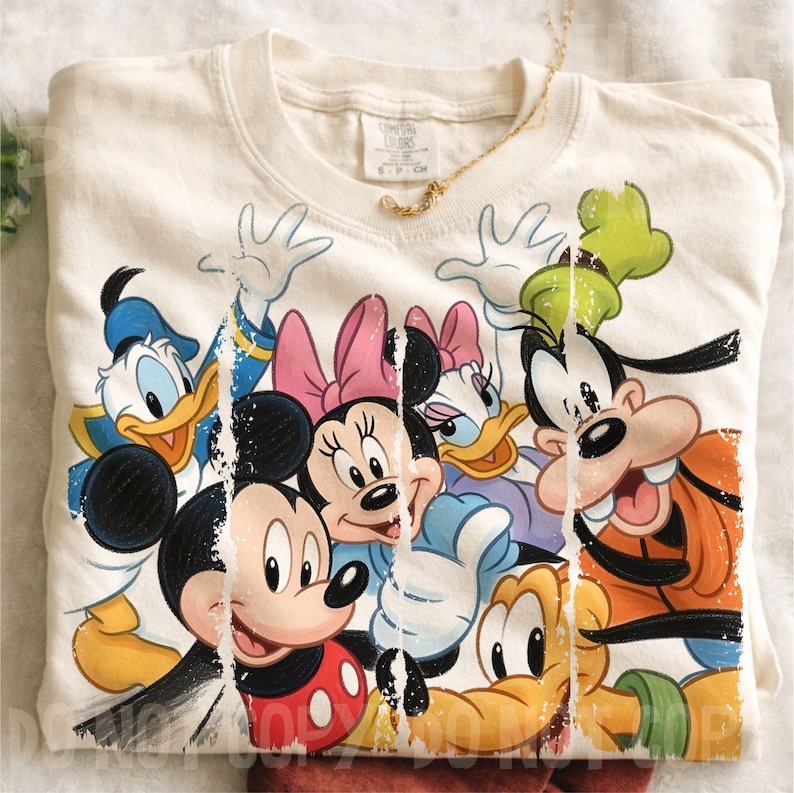 May include: Cream-colored crew neck t-shirt featuring a colorful graphic of Disney characters Mickey Mouse, Minnie Mouse, Donald Duck, Daisy Duck, Goofy, and Pluto. A gold necklace is draped across the shirt. The shirt is folded.