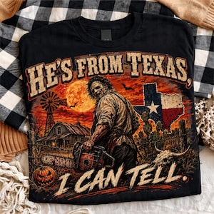 Puede incluir: Una camiseta negra con un gr&aacute;fico de una figura enmascarada sosteniendo una motosierra en un paisaje rural con un molino, granero, calabaza y la bandera de Texas. El texto dice HE'S FROM TEXAS, I CAN TELL en una fuente desgastada.