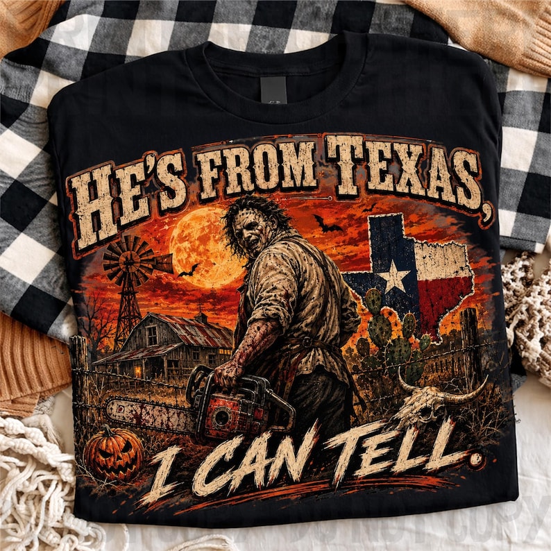 Puede incluir: Una camiseta negra con un gr&aacute;fico de una figura enmascarada sosteniendo una motosierra en un paisaje rural con un molino, granero, calabaza y la bandera de Texas. El texto dice HE'S FROM TEXAS, I CAN TELL en una fuente desgastada.