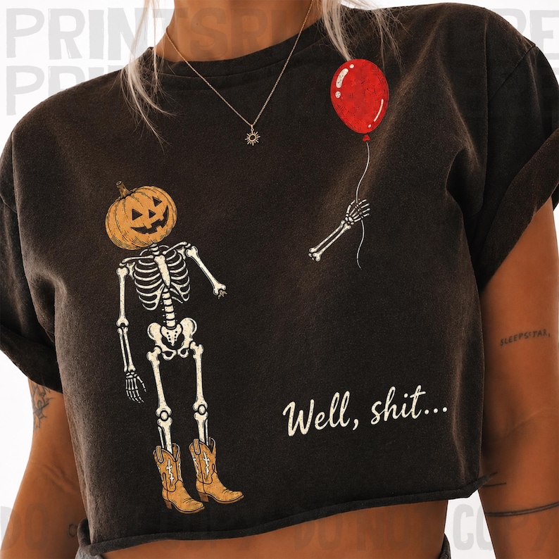 Op de afbeelding: Een donker antracietgrijs t-shirt met korte mouwen en een afbeelding van een skelet met een pompoenhoofd en bruine cowboylaarzen. Een losse skeletarm houdt een rode ballon vast. De tekst "Well, shit..." is in het wit gedrukt.