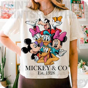 May include: Cream-colored t-shirt featuring a colorful graphic of Mickey Mouse, Minnie Mouse, Donald Duck, Daisy Duck, Goofy, and Pluto. The text "MICKEY & CO" and "Est. 1928" are printed below the characters. The shirt has short sleeves.