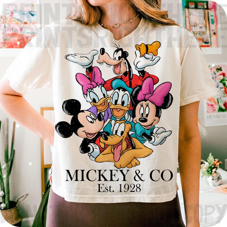 May include: Cream-colored t-shirt featuring a colorful graphic of Mickey Mouse, Minnie Mouse, Donald Duck, Daisy Duck, Goofy, and Pluto. The text "MICKEY & CO" and "Est. 1928" are printed below the characters. The shirt has short sleeves.
