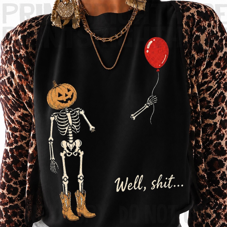 Op de afbeelding: Een zwart shirt met lange mouwen en een luipaardprint op de mouwen. Op de voorkant staat een skelet met een pompoenhoofd, bruine cowboylaarzen en een rode ballon in de hand. De tekst luidt, Well, shit...