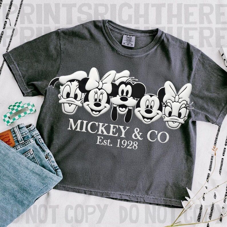 May include: Dark gray t-shirt featuring embroidered images of Mickey Mouse, Minnie Mouse, Donald Duck, and Daisy Duck. The text "Mickey & Co. Est. 1928" is embroidered below the characters. A pair of blue jeans and a hair clip are visible.