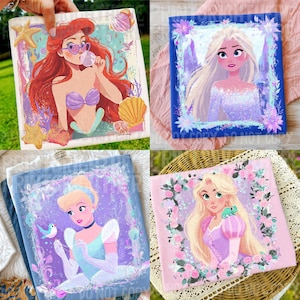 May include: Four square art prints featuring Disney princesses. Ariel with red hair and sunglasses, Elsa with blonde hair, Cinderella in a blue dress, and Rapunzel with long blonde hair. Each print has a floral border and a unique background color.
