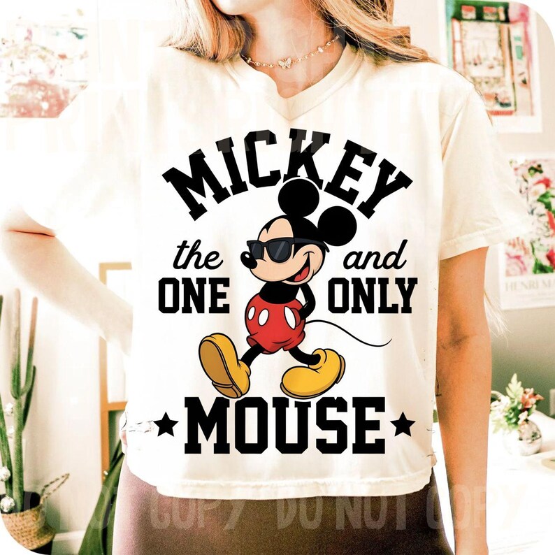 May include: Cream-coloured t-shirt featuring a graphic of Mickey Mouse wearing sunglasses, with the text "Mickey the One and Only Mouse." Mickey is depicted in his classic red shorts, yellow shoes, and black gloves. The design includes black stars.