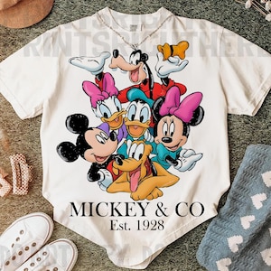 May include: Cream-colored t-shirt featuring a colorful graphic of Mickey Mouse, Minnie Mouse, Donald Duck, Daisy Duck, Goofy, and Pluto. The text "MICKEY & CO" and "Est. 1928" are printed below the characters. A silver chain necklace is visible.