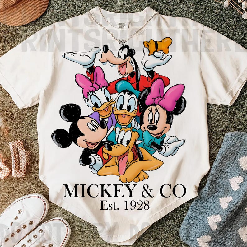 May include: Cream-colored t-shirt featuring a colorful graphic of Mickey Mouse, Minnie Mouse, Donald Duck, Daisy Duck, Goofy, and Pluto. The text "MICKEY & CO" and "Est. 1928" are printed below the characters. A silver chain necklace is visible.