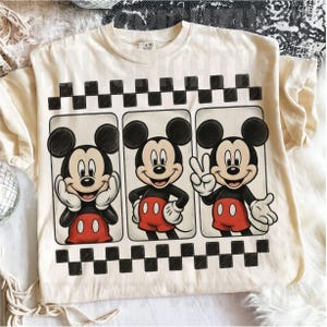 May include: Cream-colored t-shirt featuring three panels with Mickey Mouse illustrations. Each panel shows Mickey in a different pose, with a black and white checkered border above and below the panels. Mickey is wearing red shorts and white gloves.