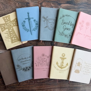 May include: A collection of nine rectangular journals in various pastel colors, each featuring unique debossed designs including a cross, sword, shield, anchor, lantern, and floral motifs, along with inspirational text like Saved by Grace.