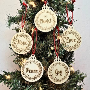 May include: Five round light-colored wooden ornaments hanging by red strings. Each ornament features a laser-engraved wreath design surrounding the words Christ, Hope, Love, Peace, or Joy in a script font, with a small star above Christ.