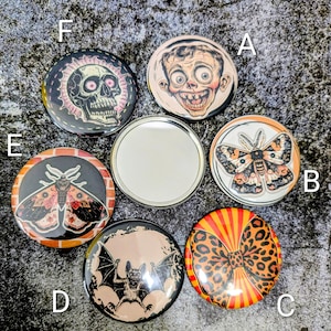 May include: A selection of circular decorative badges with diverse designs. The badges display illustrations of a skull, a moth, a bat, a bow, and a distorted face. The colour scheme includes black, orange, red, and beige.