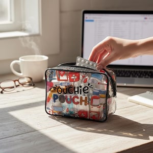 Op de afbeelding: Een doorzichtige "Ouchie Pouch" met ritssluiting, gevuld met medische benodigdheden en een kleurrijk patroon van medische symbolen. Een hand haalt een blisterverpakking met pillen uit de pouch. Een kop koffie, een bril en een laptop staan op de achtergrond.