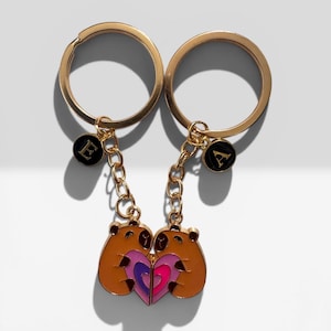 Puede incluir: Dos llaveros dorados con un diseño de oso marrón abrazando un corazón en rosa, morado y azul. Cada llavero tiene un anillo dorado y una cadena con un dije negro con una letra. El fondo es blanco.