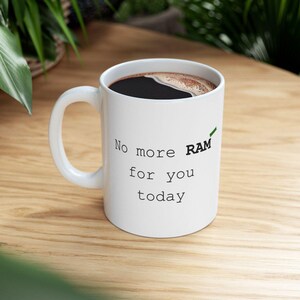 May include: White ceramic coffee mug with the text "No more RAM for you today" in black, with the word "RAM" in green. The mug is filled with dark coffee and sits on a wooden surface.