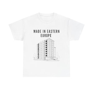 Può includere: T-shirt bianca con la scritta "MADE IN EASTERN EUROPE" sopra un'illustrazione in bianco e nero di un edificio a più piani. La grafica raffigura un edificio con molte finestre e un tetto piatto, con alberi davanti.