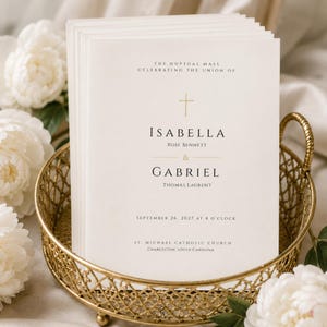 May include: A stack of off-white wedding program cards featuring gold foil text and a cross symbol. The text reads: The Nuptial Mass Celebrating the Union of Isabella Rose Bennett & Gabriel Thomas Laurent, September 26, 2027 at 4 o'clock, St. Michael Catholic Church, Charleston, South Carolina.