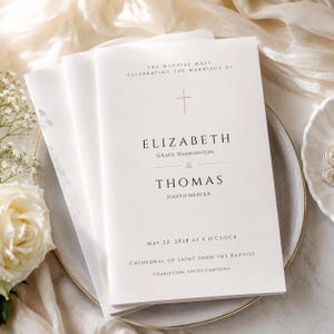 May include: A white wedding program booklet featuring a gold cross icon and black serif text that reads: The Nuptial Mass celebrating the marriage of Elizabeth Grace Harrington and Thomas Joseph Mercer, May 22, 2028 at 4 o'clock, Cathedral of Saint John the Baptist, Charleston, South Carolina.