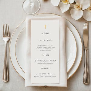May include: A formal table setting with a white plate, gold rim, and a menu card. The menu card has a gold cross and lists a first course, entree, and dessert. A fork and knife are on either side of the plate.