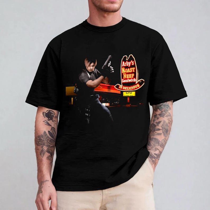 May include: Black t-shirt featuring a graphic of a character holding a gun in front of an Arby's sign. The sign reads "Arby's Roast Beef Sandwich is Delicious" in red and yellow neon. The character is in a black outfit.