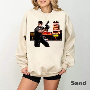 May include: Sand-colored sweatshirt featuring a graphic of a person holding a gun in front of an Arby's restaurant sign. The sign reads "Arby's Roast Beef Sandwich Is Delicious" in red and yellow. The sweatshirt is a neutral color.