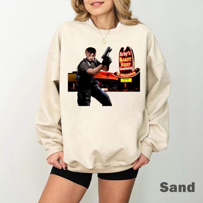 May include: Sand-colored sweatshirt featuring a graphic of a person holding a gun in front of an Arby's restaurant sign. The sign reads "Arby's Roast Beef Sandwich Is Delicious" in red and yellow. The sweatshirt is a neutral color.
