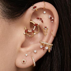 Könnte beinhalten: Eine Sammlung goldfarbener Ohrschmuckstücke für mehrere Piercings. Das Set umfasst verschiedene Creolen, Stecker und hängende Anhänger, die mit kleinen klaren weißen Kristallen und tiefroten Edelsteinen im Marquise-Schliff verziert sind.