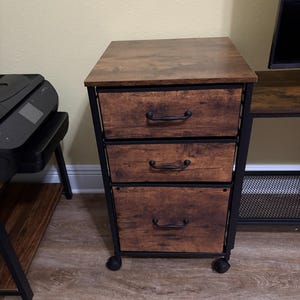 May include: A three-drawer wooden file cabinet with a dark brown finish and black metal frame. The cabinet sits on small black wheels and features curved black handles. The top of the cabinet is also dark brown.
