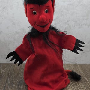 May include: A plush devil doll with a red body, face, and horns. The doll has black hair and a tail. The doll's arms and hands are black. The doll is sitting on a wooden surface.