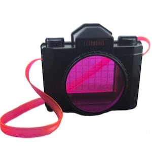 Peut inclure: Un appareil photo jouet en plastique noir avec le mot LEADWORKS imprimé en petites lettres sur le devant. L'appareil possède un grand objectif circulaire doté d'un filtre translucide rose vif avec un motif quadrillé et une longue sangle rose néon.