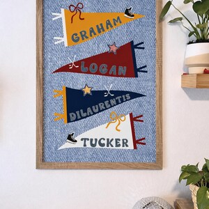 May include: A framed wall art piece featuring four pennant flags with names: Graham, Logan, Dilauentis, and Tucker. Each pennant is a different color and decorated with sports-themed icons. The background is a blue denim print.