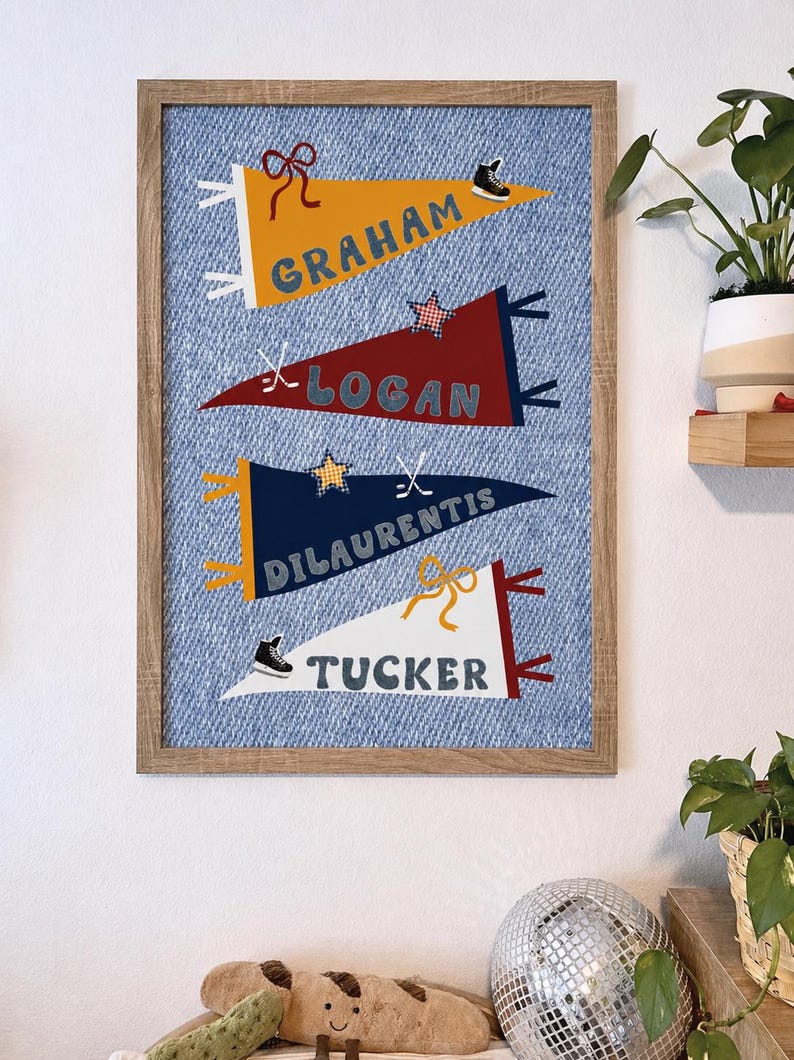 May include: A framed wall art piece featuring four pennant flags with names: Graham, Logan, Dilauentis, and Tucker. Each pennant is a different color and decorated with sports-themed icons. The background is a blue denim print.