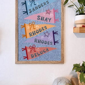 May include: A framed print featuring pennant flags with names and numbers. The flags are in various colors, including blue, pink, orange, and purple, set against a denim-textured background. Each pennant has a bow and sports-themed icons. The frame is made of light-colored wood.