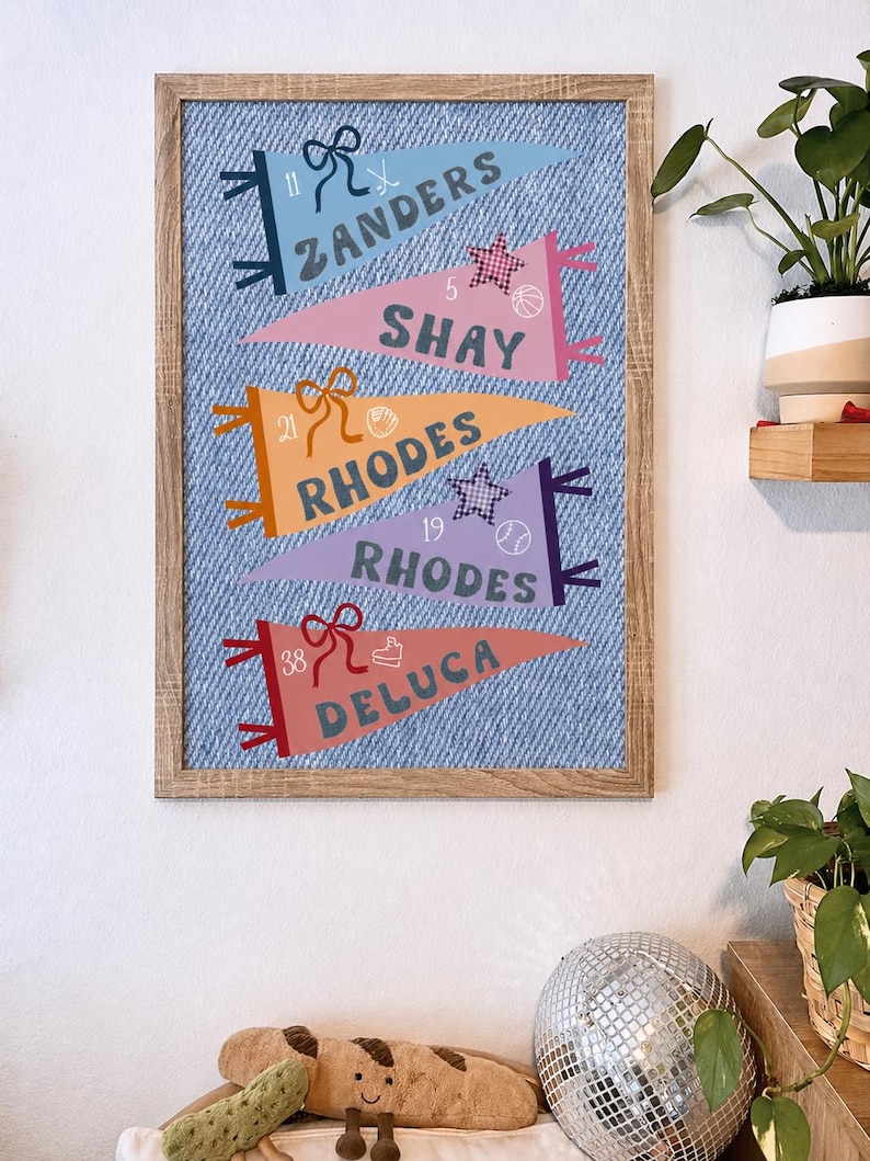 May include: A framed print featuring pennant flags with names and numbers. The flags are in various colors, including blue, pink, orange, and purple, set against a denim-textured background. Each pennant has a bow and sports-themed icons. The frame is made of light-colored wood.