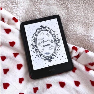 May include: A black Kindle e-reader displaying a decorative design with the text "alyssa's kindle." The e-reader is on a white blanket with red heart patterns. The Kindle is a portable device for reading.