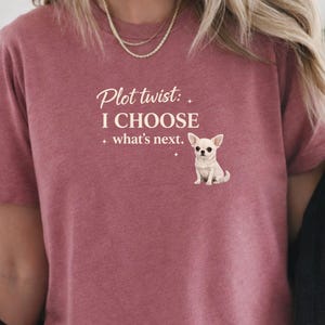 Puede incluir: Una camiseta de manga corta color malva con el texto impreso en blanco Plot twist: I CHOOSE what's next. acompañado de un pequeño gráfico de un perro chihuahua blanco. La camiseta tiene cuello redondo y una textura jaspeada en la tela.