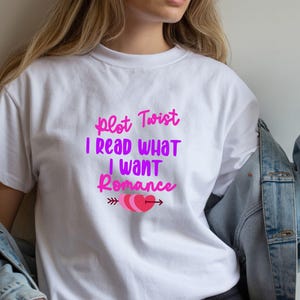 May include: A white short-sleeved t-shirt featuring the pink and purple text "Plot Twist I READ WHAT I WANT Romance" printed on the chest, accompanied by a graphic of three overlapping pink hearts pierced by a horizontal arrow.