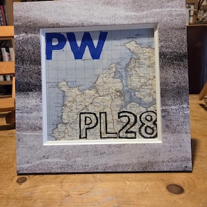 Puede incluir: Impresión de mapa enmarcada con un borde moteado gris y blanco. El mapa presenta un diseño geográfico detallado con las letras azules "PW" en la parte superior y las letras y números negros "PL28" en la parte inferior.